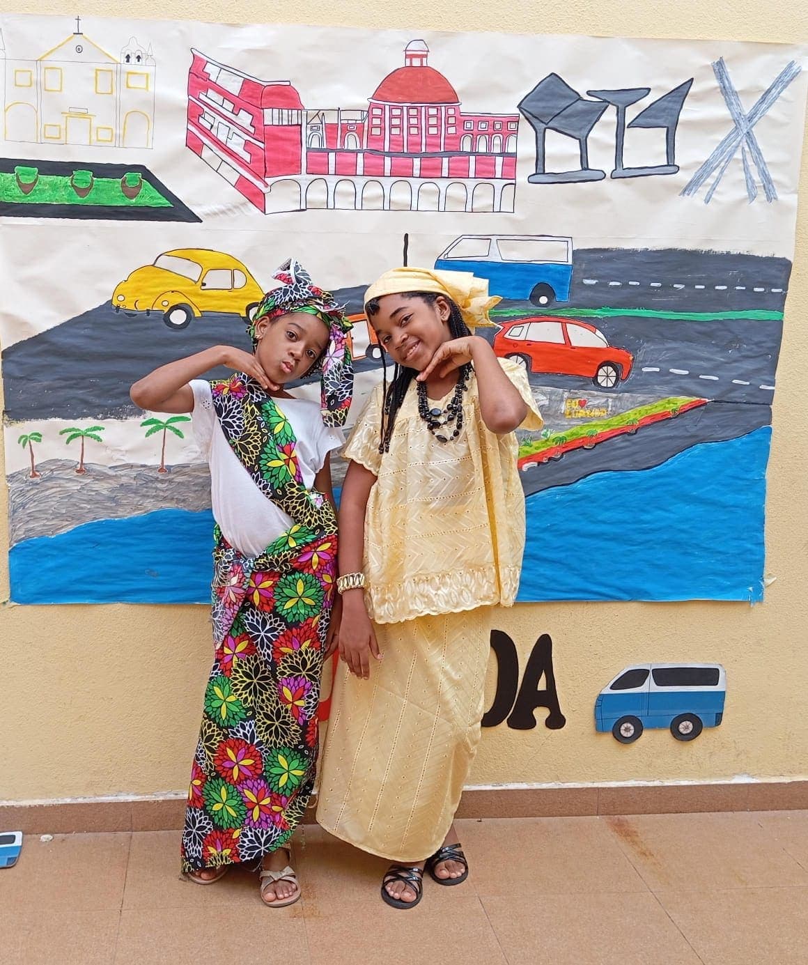 Duas alunas em traje tradicional, em frente ao cenário de Luanda
