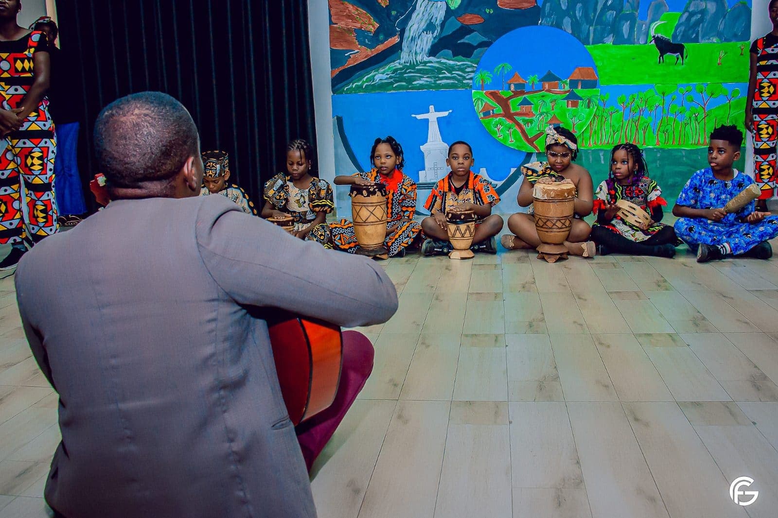 O professor à viola, com os alunos sentados em frente ao mural